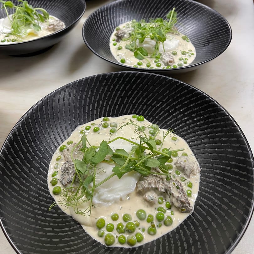 Three black bowls of creamy risotto with green peas, morel mushrooms, poached eggs, and pea shoots.