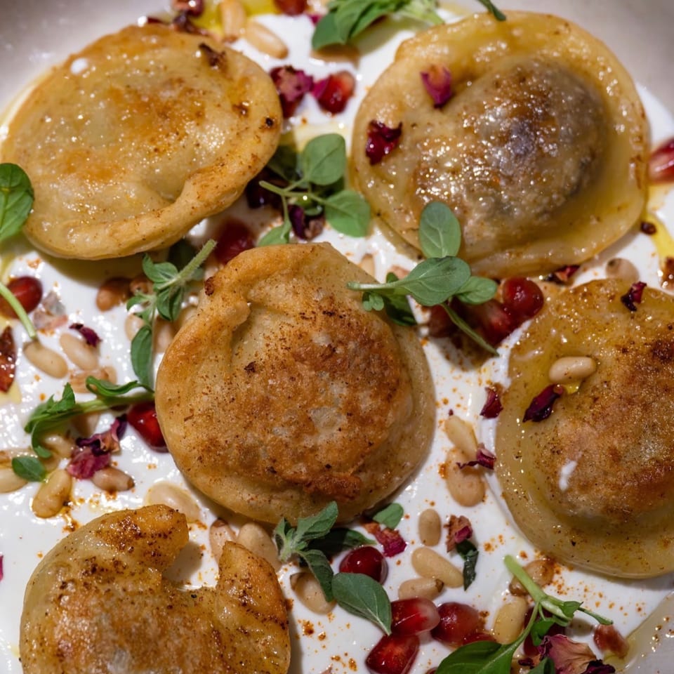 Five golden-brown dumplings on a white plate with yogurt, pine nuts, pomegranate seeds, and herbs.