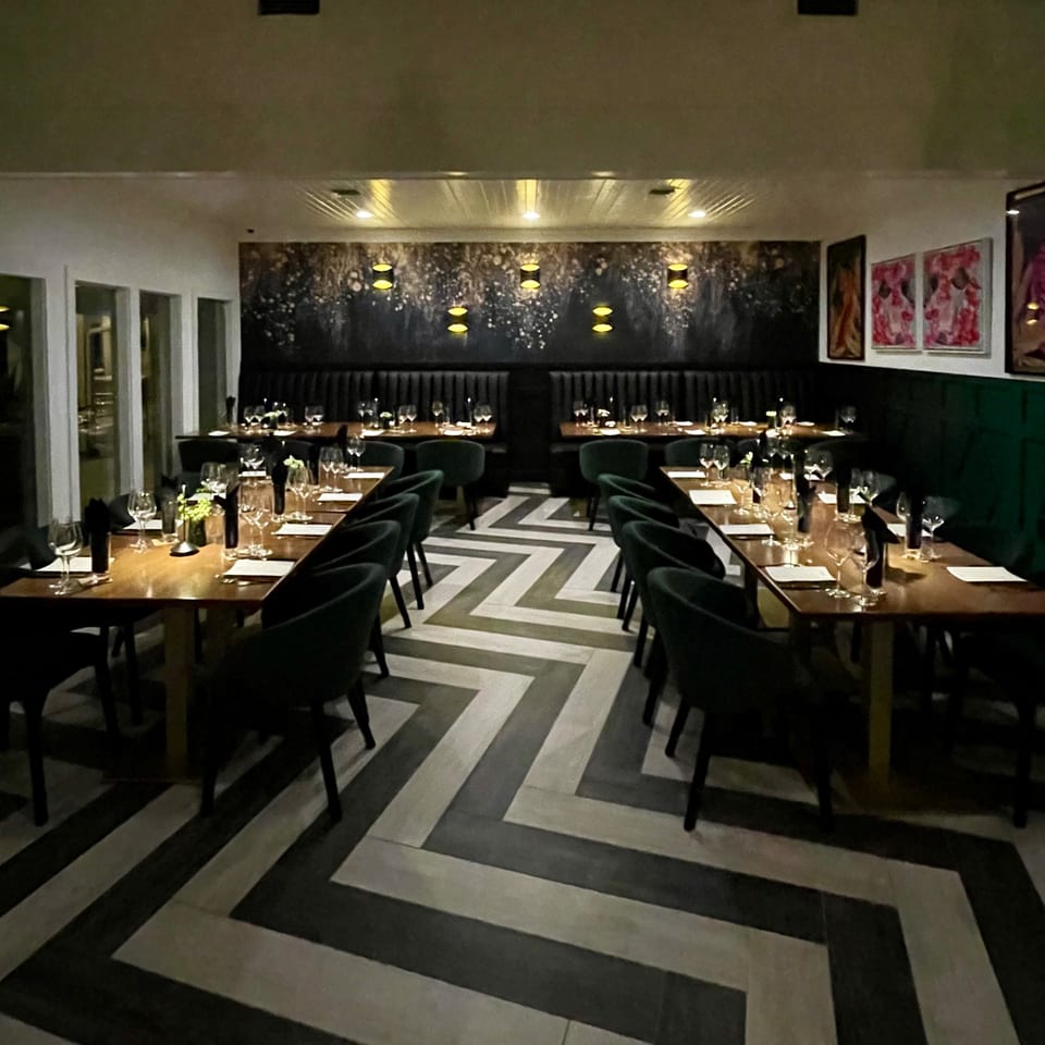 A dimly lit restaurant dining room with set tables, dark chairs, and a chevron-patterned floor.