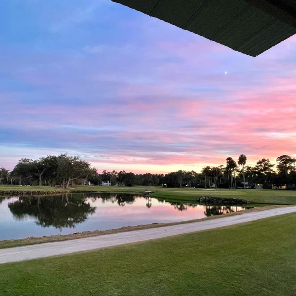 A vibrant pink and blue sunset sky over a golf course with a reflective pond and trees.