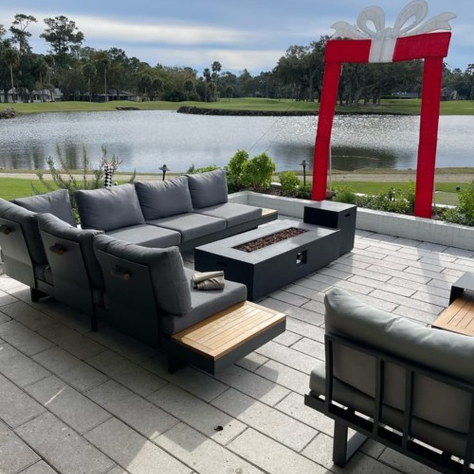 Outdoor patio with gray sectional seating, a fire pit table, and a large red gift box decoration by a lake.