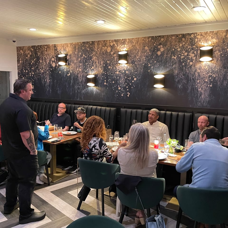 A man in a black shirt stands talking to a group of people seated at a restaurant table.