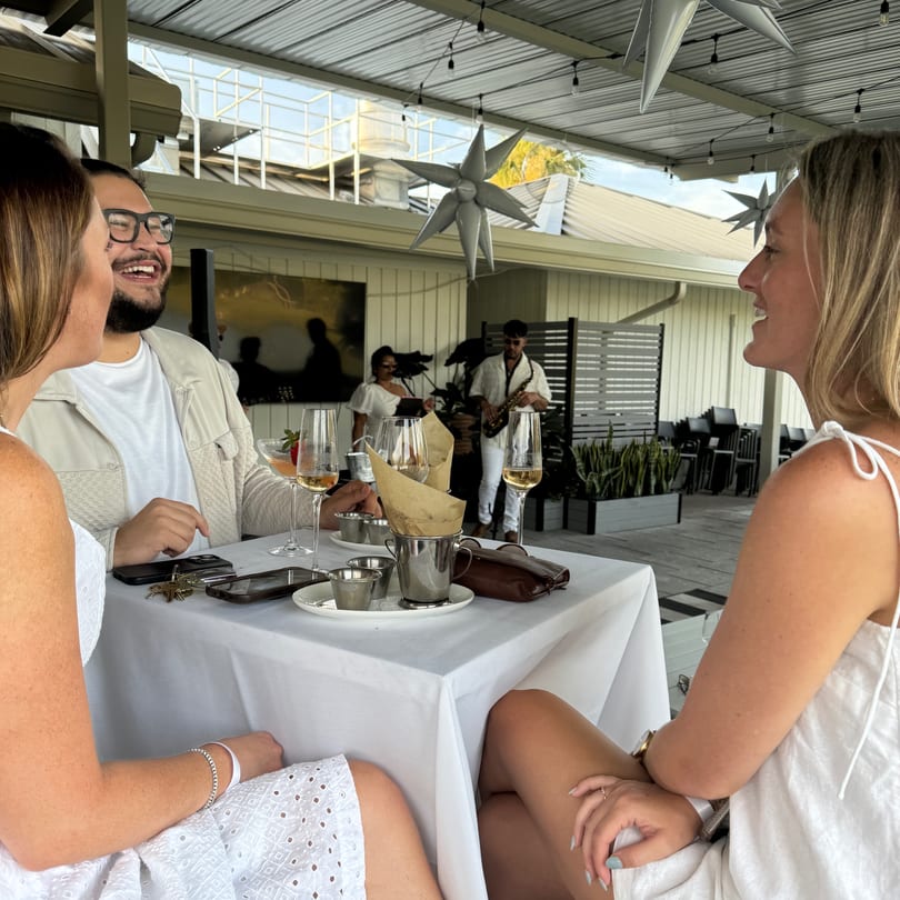 Three people enjoying drinks at an outdoor restaurant table with live music in the background.
