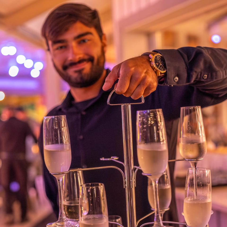 Smiling bartender holding a metal tray with multiple champagne flutes filled with bubbly drinks.