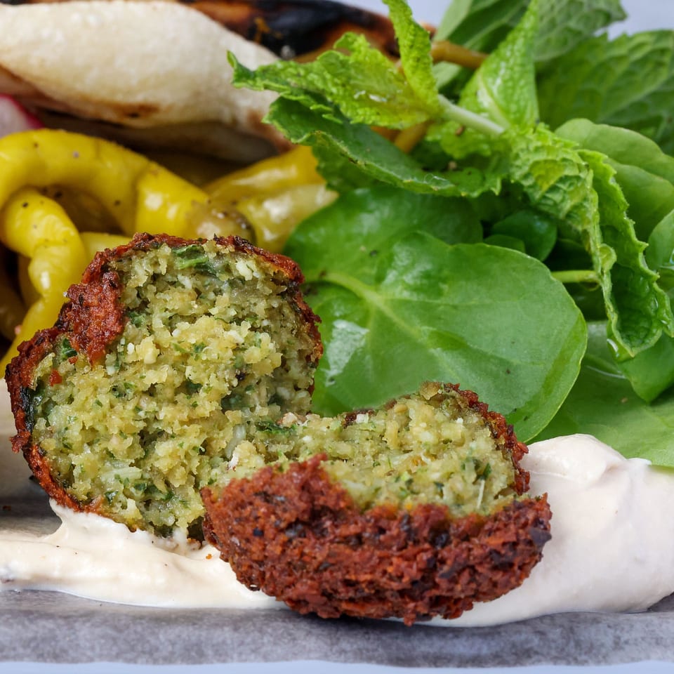 Close-up of a falafel cut in half, revealing its green interior, with sauce, peppers, and greens.