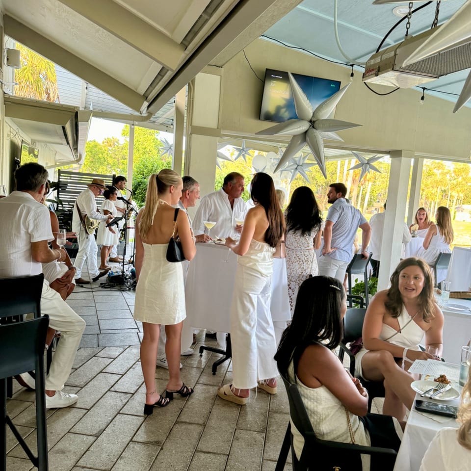 Guests in white attire mingle at an outdoor restaurant event with a live band.