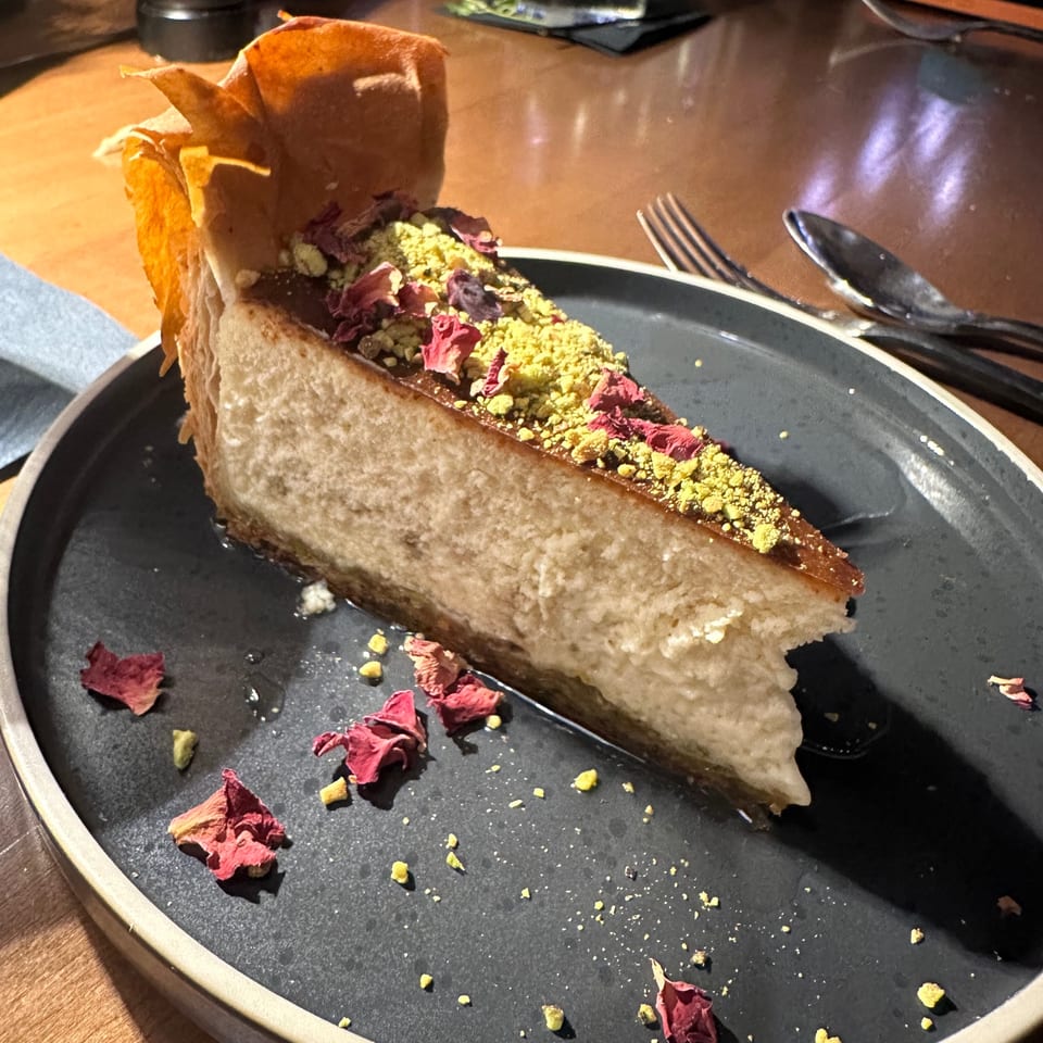 A slice of cheesecake topped with pistachios and rose petals on a dark plate.